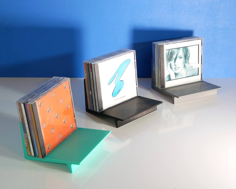 May include: Three CD holders in teal, black, and silver, each holding a stack of CDs. The teal holder is on the left, the black in the middle, and the silver on the right. The CDs have various album art, including a blue design and a portrait.
