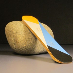 May include: A small, colorful snowboard with a geometric design. The board features sections of yellow, white, blue, and black. It rests against a large, gray stone, showcasing its design and shape.