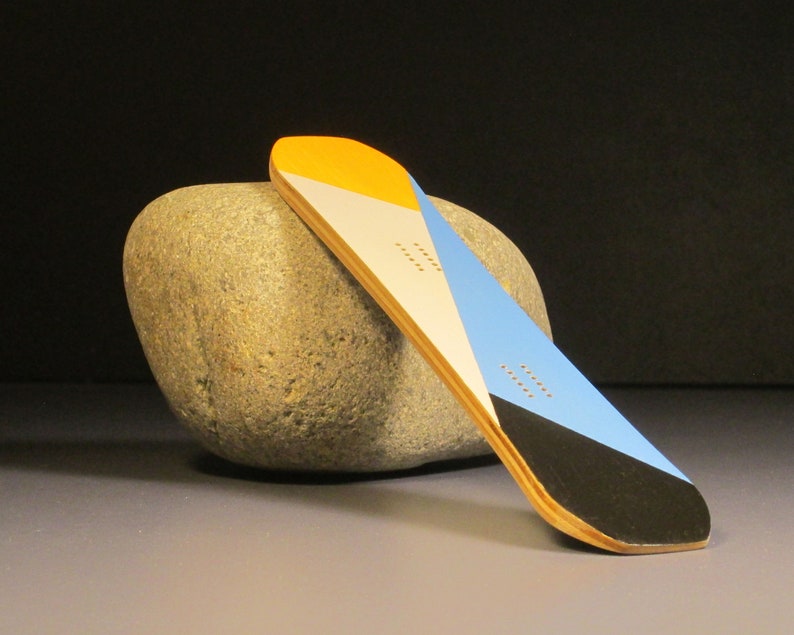 May include: A small, colorful snowboard with a geometric design. The board features sections of yellow, white, blue, and black. It rests against a large, gray stone, showcasing its design and shape.