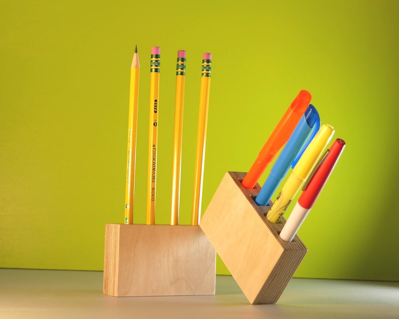 May include: A wooden desk organizer with yellow pencils and colorful markers. The pencils are yellow with pink erasers. The markers are orange, blue, yellow, and red. The organizer is made of light-colored wood.