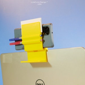 May include: A yellow 3D-printed laptop accessory with a phone holder, pen holders, and a notepad slot. The accessory is attached to a laptop, with a phone and pens in place. The text "LineColorDesign USA" is visible.
