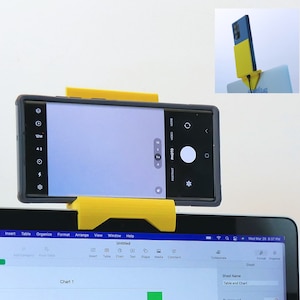 May include: A yellow smartphone holder is attached to a laptop, with a black smartphone in place. The phone's camera is facing forward. The laptop screen displays a spreadsheet. A smaller image shows the phone holder and phone.