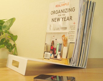 May include: A white metal magazine rack holding a copy of "Real Simple Organizing for the New Year." A smartphone rests on the wooden surface in front of the rack. A green plant is visible on the left.