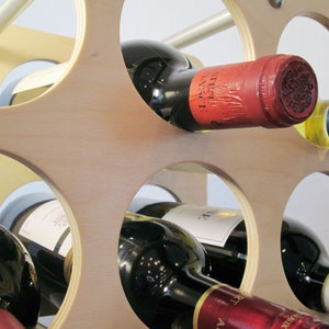 May include: A wooden wine rack with multiple circular openings, holding several wine bottles. The bottles are various colors, with red, white, and blue labels. The rack is made of light-colored wood and has a modern design.