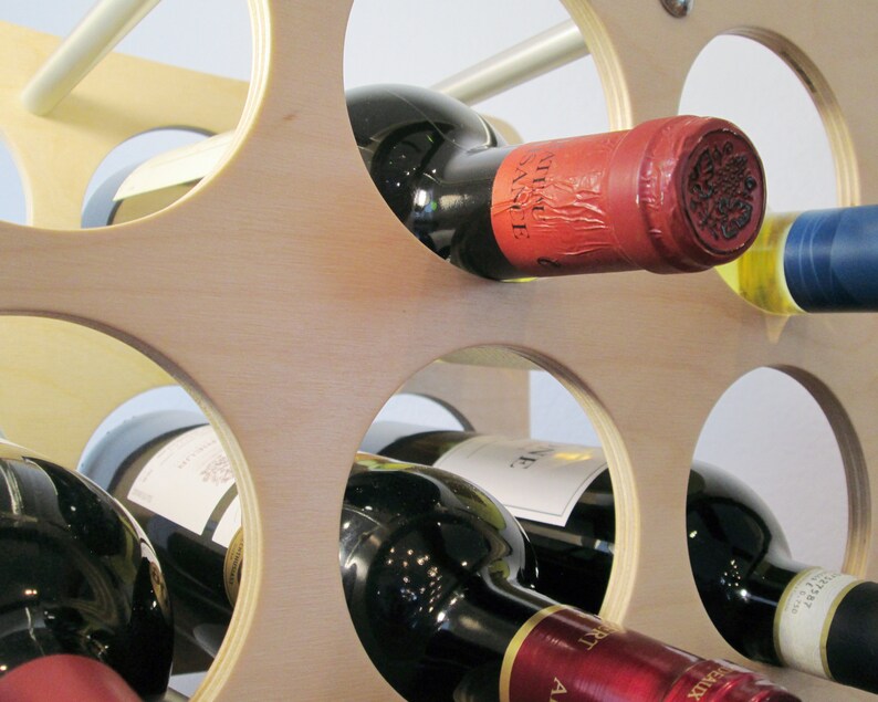 May include: A wooden wine rack with multiple circular openings, holding several wine bottles. The bottles are various colors, with red, white, and blue labels. The rack is made of light-colored wood and has a modern design.