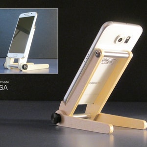 May include: A handmade wooden phone stand with a white smartphone resting on it. The stand is made of light-colored wood and has a simple, adjustable design. The words "Handmade USA" are visible.