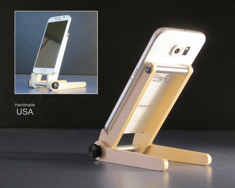 May include: A handmade wooden phone stand with a white smartphone resting on it. The stand is made of light-colored wood and has a simple, adjustable design. The words "Handmade USA" are visible.