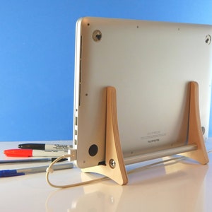 May include: A silver MacBook Pro laptop is supported by a light wood stand. The stand has two upright supports and a horizontal bar. A white charging cable is plugged in. Several pens and a marker are in the foreground.