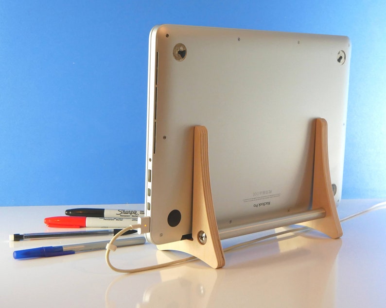 May include: A silver MacBook Pro laptop is supported by a light wood stand. The stand has two upright supports and a horizontal bar. A white charging cable is plugged in. Several pens and a marker are in the foreground.