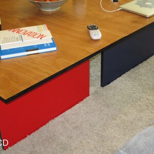 May include: A modern coffee table with a light wood grain top and contrasting red and navy blue supports. The table has a rectangular shape and is placed on a light-colored carpet. Books and a remote are visible on the table.