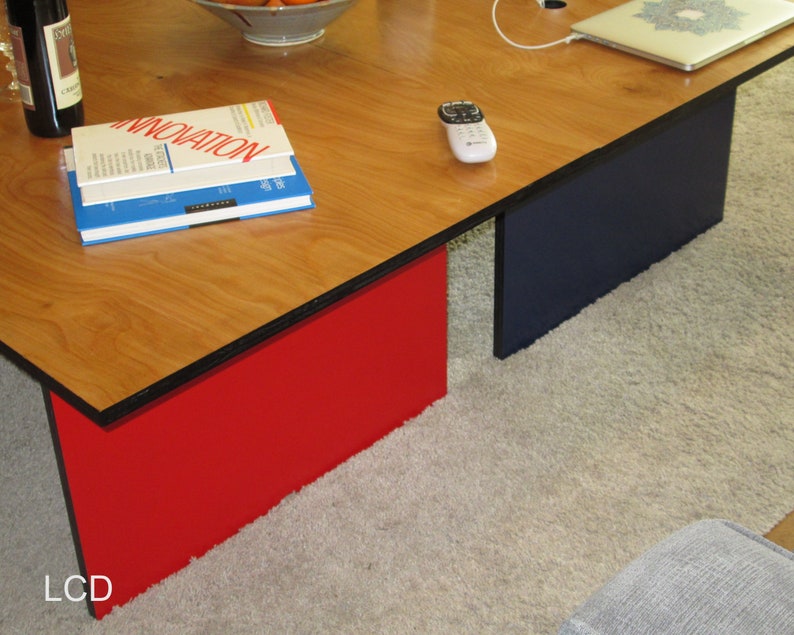 May include: A modern coffee table with a light wood grain top and contrasting red and navy blue supports. The table has a rectangular shape and is placed on a light-colored carpet. Books and a remote are visible on the table.