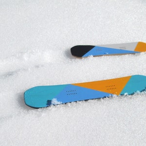 May include: Two snowboards with colorful geometric designs rest on a snowy surface. The boards feature blue, orange, black, and gray sections. One board is larger than the other, suggesting different sizes for snowboarding.