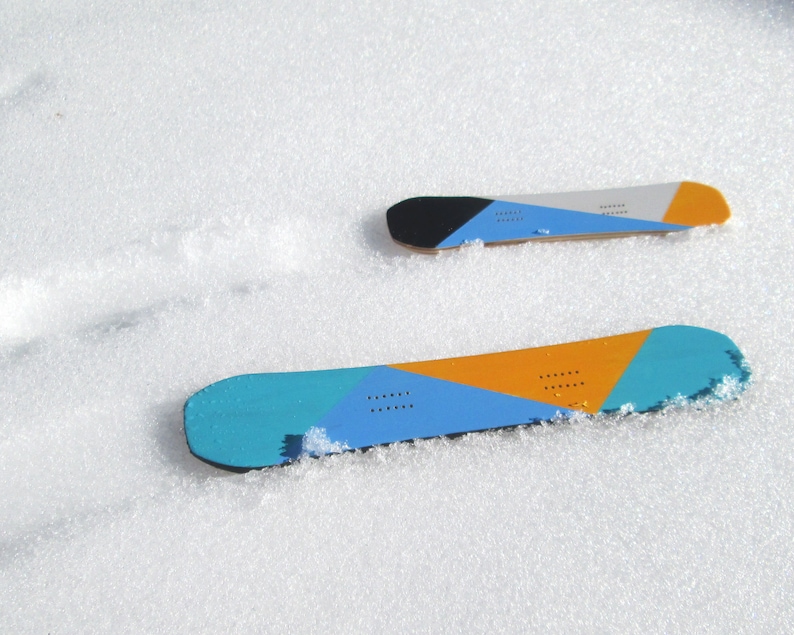 May include: Two snowboards with colorful geometric designs rest on a snowy surface. The boards feature blue, orange, black, and gray sections. One board is larger than the other, suggesting different sizes for snowboarding.