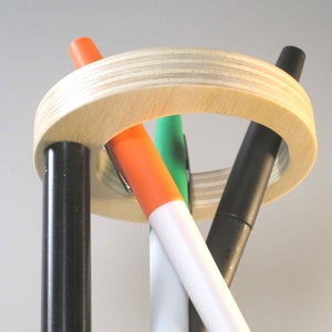 May include: A wooden pen holder with a light wood finish, holding four colorful markers. The markers are black, orange, green, and white. The holder has a circular design with vertical supports.