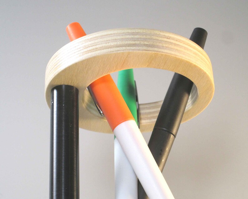 May include: A wooden pen holder with a light wood finish, holding four colorful markers. The markers are black, orange, green, and white. The holder has a circular design with vertical supports.
