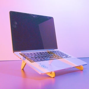 May include: A silver laptop computer is elevated on a light-colored wooden stand. The laptop screen is black, and the keyboard is visible. The stand has a simple design with two angled legs, providing an ergonomic setup for the device.