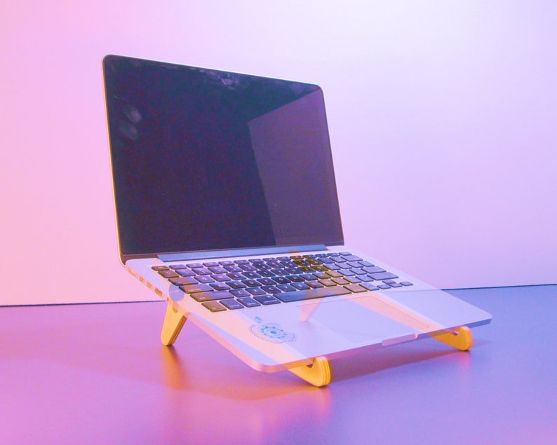 May include: A silver laptop computer is elevated on a light-colored wooden stand. The laptop screen is black, and the keyboard is visible. The stand has a simple design with two angled legs, providing an ergonomic setup for the device.