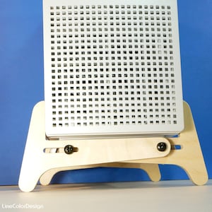 May include: A white, square, perforated panel is mounted on a light wood easel. The easel has a two-piece design with adjustable angles, secured by black screws. The background is a solid blue.