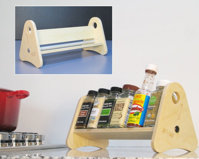 May include: A light-colored wooden spice rack with a triangular frame and a shelf for holding spice jars. The rack is displayed on a kitchen counter, holding various spice bottles. The rack has a height of approximately 8 inches.