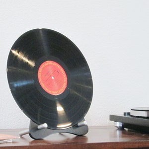 Op de afbeelding: Een zwarte vinylplaat met een rood-wit label wordt op een zwarte standaard gepresenteerd. De plaat staat schuin, waardoor het oppervlak zichtbaar is. Op de achtergrond is een platenspeler te zien, wat wijst op een muzikaal thema.