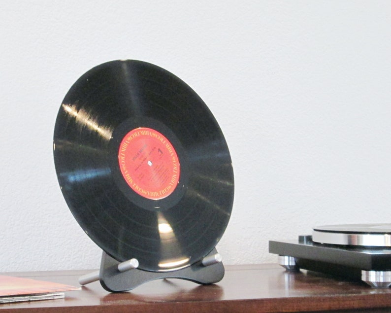 May include: A black vinyl record with a red and white label is displayed on a black stand. The record is angled, showcasing its surface. A turntable is visible in the background, suggesting a music theme.