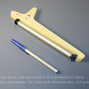May include: A wooden and metal device with a pen. The device has a light wood frame and a silver metal roller. The pen is blue and silver. The text in the image says, "Folds down nice and small to fit a backpack or laptop cases and sleeves. No Tools and ultralight 5oz, 145 grams."