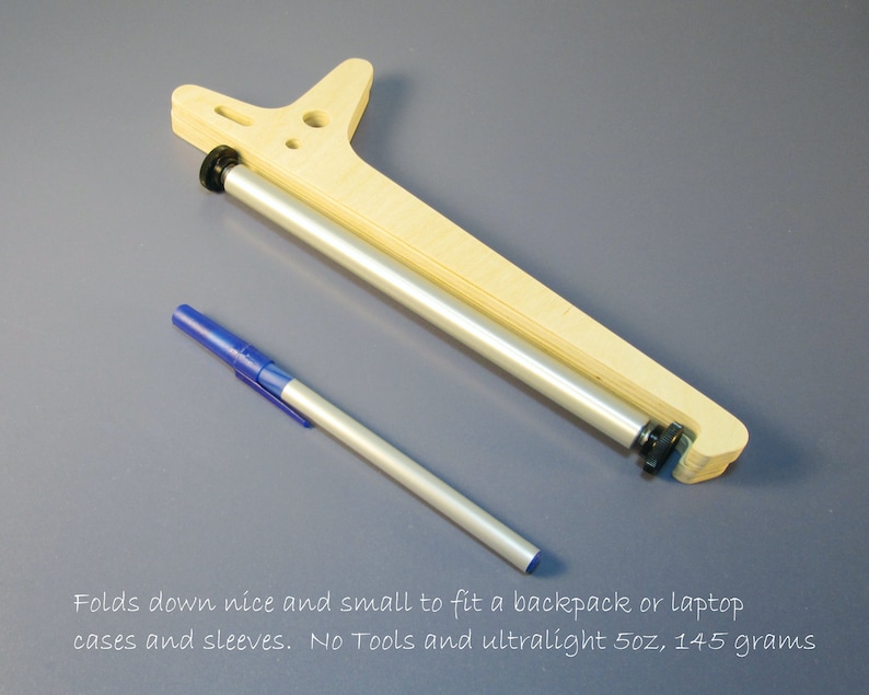 May include: A wooden and metal device with a pen. The device has a light wood frame and a silver metal roller. The pen is blue and silver. The text in the image says, "Folds down nice and small to fit a backpack or laptop cases and sleeves. No Tools and ultralight 5oz, 145 grams."