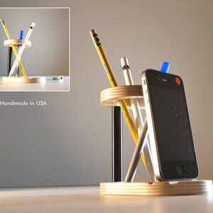 May include: A wooden desk organizer with a circular design, holding pencils, pens, and a smartphone. The organizer is made of light-colored wood and has a minimalist aesthetic. The text "Handmade in USA" is visible.