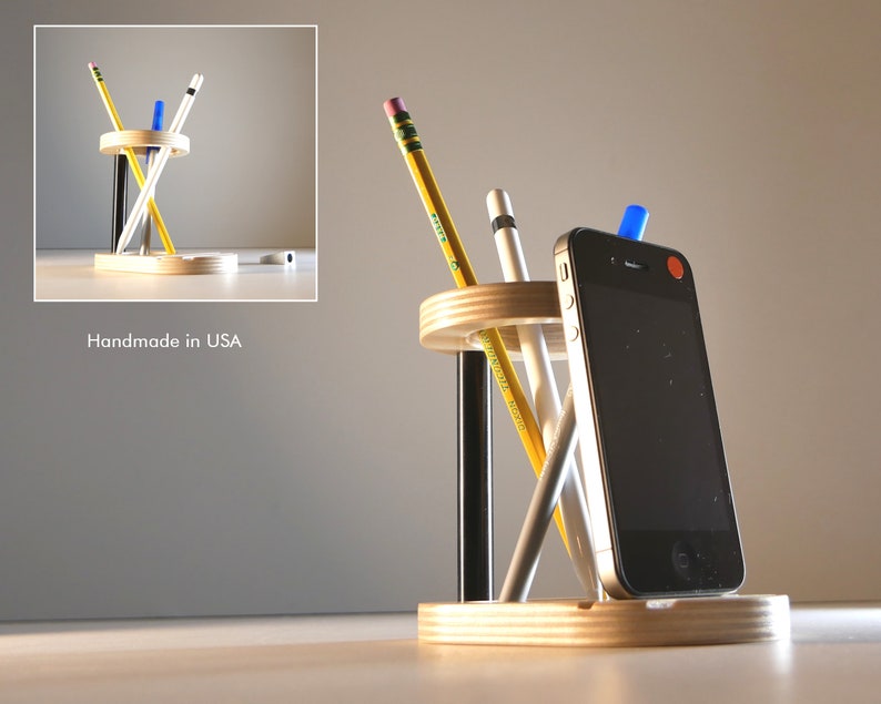 May include: A wooden desk organizer with a circular design, holding pencils, pens, and a smartphone. The organizer is made of light-colored wood and has a minimalist aesthetic. The text "Handmade in USA" is visible.