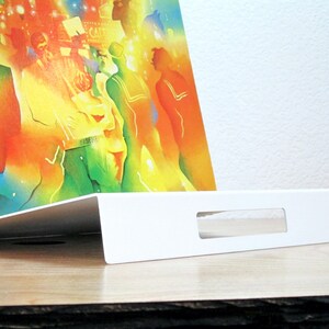 May include: A white, angled display stand with a rectangular cutout, holding a colorful print. The print features a vibrant, abstract scene with figures in various colors. The stand is on a wooden surface.