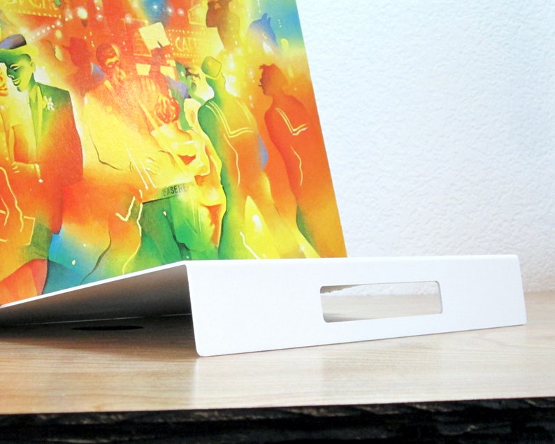May include: A white, angled display stand with a rectangular cutout, holding a colorful print. The print features a vibrant, abstract scene with figures in various colors. The stand is on a wooden surface.