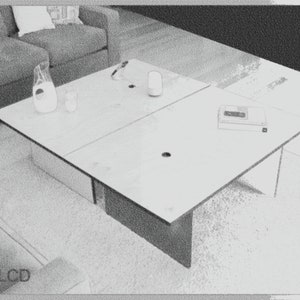 May include: A light-colored, modern coffee table with a rectangular top, divided into four sections. A pitcher, two glasses, and a small electronic device are on the table. The table is in front of a gray sofa.