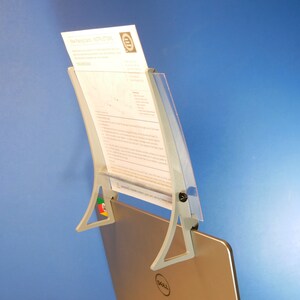 Op de afbeelding: Een grijze documenthouder bevestigd aan een laptop. De houder heeft een gebogen ontwerp met een doorzichtige plastic voorkant en een metalen basis. Een document wordt op zijn plaats gehouden. De laptop heeft een Dell-logo.