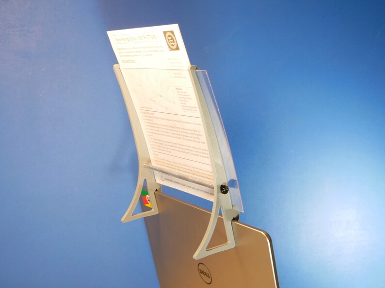 Op de afbeelding: Een grijze documenthouder bevestigd aan een laptop. De houder heeft een gebogen ontwerp met een doorzichtige plastic voorkant en een metalen basis. Een document wordt op zijn plaats gehouden. De laptop heeft een Dell-logo.