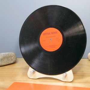 May include: A black vinyl record with an orange label that reads "Guitar Magic Play Along Records" is displayed on a light wood stand. The record is centered, showcasing its grooves. The stand has a minimalist design with silver metal supports.