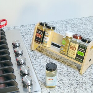 May include: A wooden spice rack holding several spice jars, including "Garlic", "Onion Salt", and "Rosemary". The rack sits on a countertop next to a stainless steel stovetop and a red pot. A single spice jar sits in front of the rack.