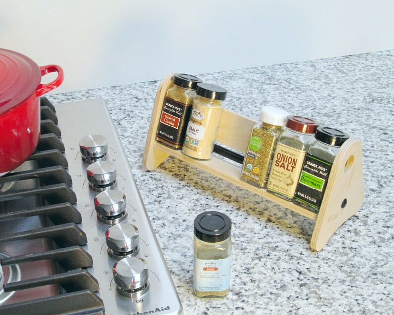 May include: A wooden spice rack holding several spice jars, including "Garlic", "Onion Salt", and "Rosemary". The rack sits on a countertop next to a stainless steel stovetop and a red pot. A single spice jar sits in front of the rack.