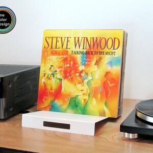 A modern white sheet metal stand for storing vinyl records leaning back at a small angle.  Shown here on a console between a turntable and a stereo receiver. Minimalist styling for a very modern touch.  A simple design with two ninety degree bends.