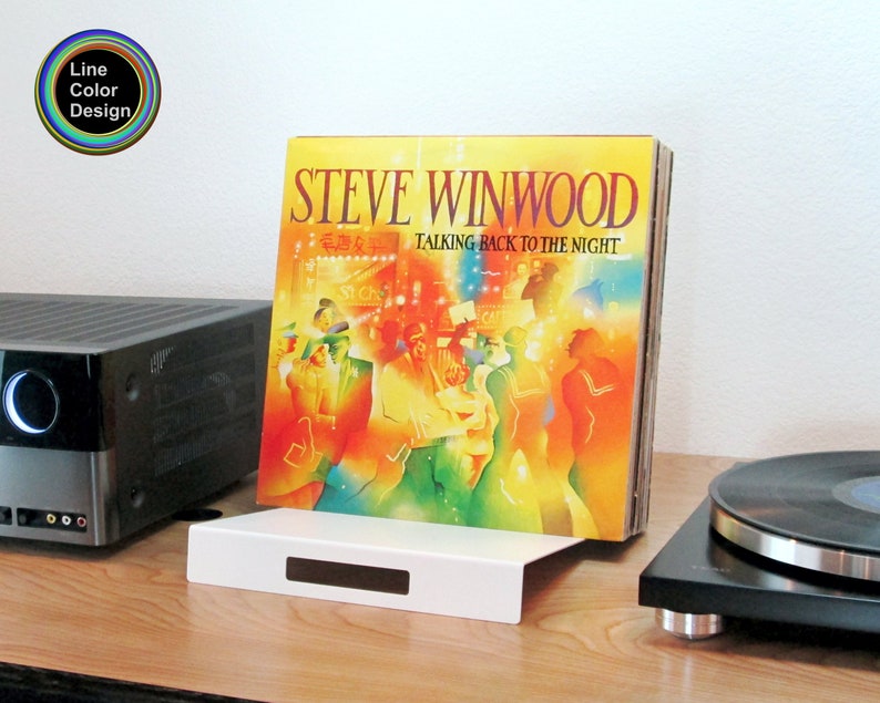 A modern white sheet metal stand for storing vinyl records leaning back at a small angle.  Shown here on a console between a turntable and a stereo receiver. Minimalist styling for a very modern touch.  A simple design with two ninety degree bends.