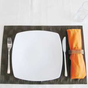 May include: A place setting with a white square plate, a fork, and a knife on a brown placemat. An orange napkin is held by a wooden napkin ring. A clear glass is in the background.