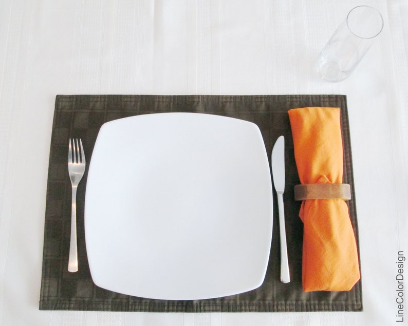 May include: A place setting with a white square plate, a fork, and a knife on a brown placemat. An orange napkin is held by a wooden napkin ring. A clear glass is in the background.