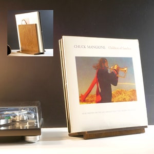 May include: A wooden record album display stand holding a vinyl record album. The album cover features a man playing a trumpet with a red scarf against a desert landscape. The album is titled "Children of Sanchez" by Chuck Mangione.