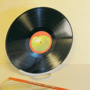 May include: A black vinyl record with a red and yellow label, displayed on a white stand. The record is tilted, showcasing its grooves and the label's design. The album cover, with text, is partially visible.