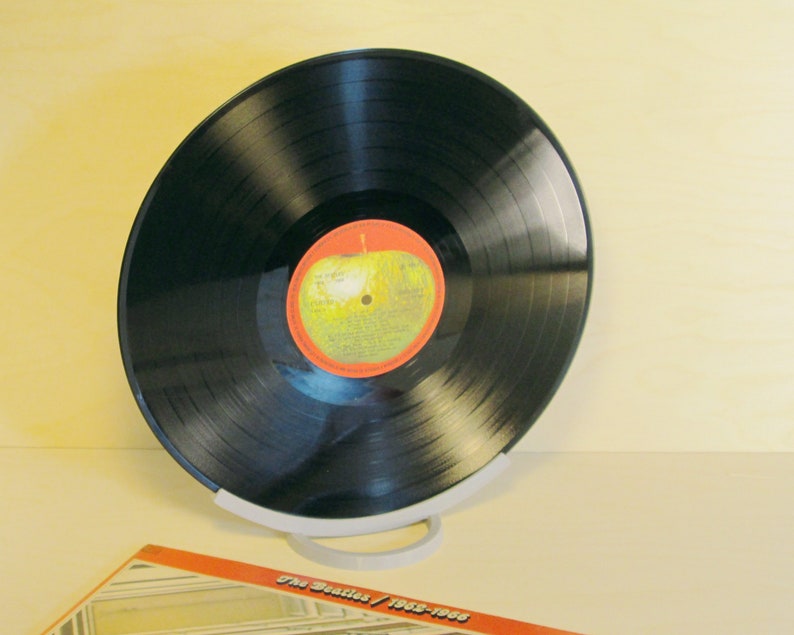 May include: A black vinyl record with a red and yellow label, displayed on a white stand. The record is tilted, showcasing its grooves and the label's design. The album cover, with text, is partially visible.