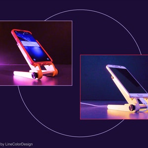 May include: A wooden phone stand with a white and orange design, holding a smartphone. The stand has a rectangular base and a slanted support for the phone. The phone is charging with a white cable. The design is by LineColorDesign.