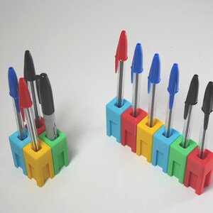 May include: Two pen holders with pens. One holder is a square of four connected blocks, and the other is a row of six connected blocks. The holders are made of colorful plastic blocks in red, yellow, green, and blue. The pens are red, blue, and black.