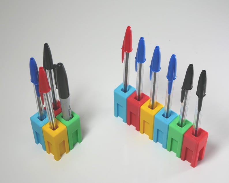 May include: Two pen holders with pens. One holder is a square of four connected blocks, and the other is a row of six connected blocks. The holders are made of colorful plastic blocks in red, yellow, green, and blue. The pens are red, blue, and black.