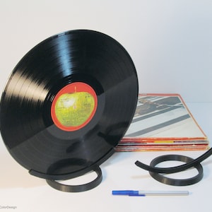 May include: A black vinyl record displayed in a black stand. The record has a red and yellow label. Additional black stands and a stack of album covers are visible. A blue pen is also present.