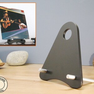 May include: A dark gray metal record display stand with a triangular shape and a circular hole at the top. The stand holds a vinyl record album with a photo of two guitar players. The stand has two silver pegs.