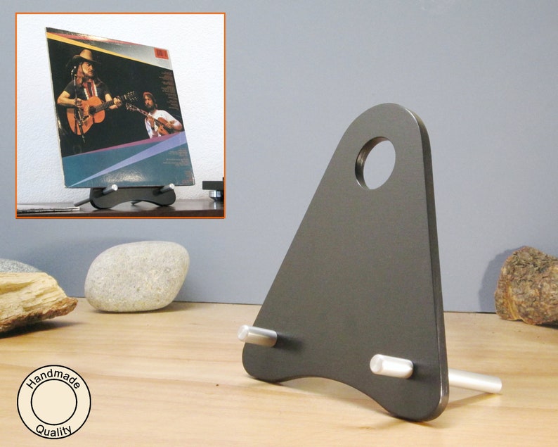 May include: A dark gray metal record display stand with a triangular shape and a circular hole at the top. The stand holds a vinyl record album with a photo of two guitar players. The stand has two silver pegs.